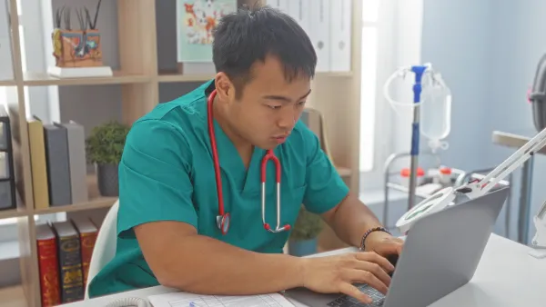 asian man in green scrubs typing into a computer at a desk. He has a stethoscope around his neck and he's in a well-lit room with a bookshelf behind him and other medical equipment.