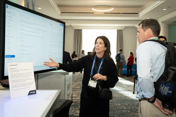 a woman in black pointing to a large monitor with a EHR demo. there is a man with a backpack watching on the right side of the screen. they are standing in the foyer a hotel ballroom