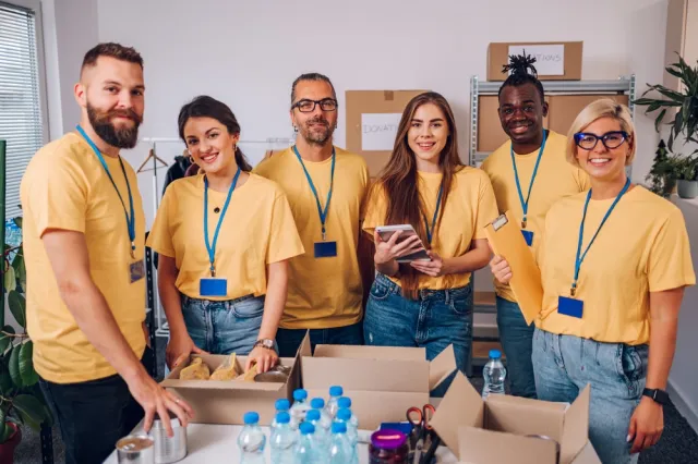 6 people in yellow shirts with blue badges in a room. they are a diverse group of people and they are packing cardboard boxes with water bottles and supplies. they're all looking at the camera and smiling