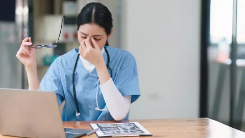 female doctor in blue scrubs holding her nose and glasses in another hand with a computer on the desk in front of her