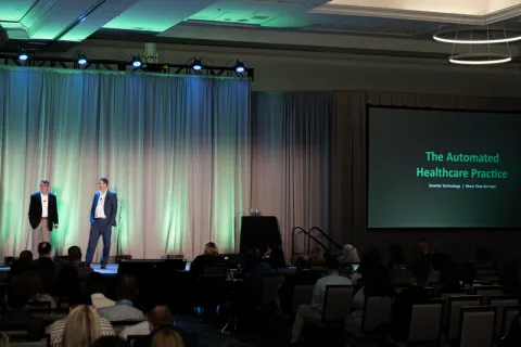 2 men standing on stage with green lights behind them, a screen to their left with "The Automated Healthcare Practice" printed
