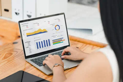 a laptop sitting on a wood desk. A woman is typing on the keyboard, her back is to the camera. The screen shows charts and graphs