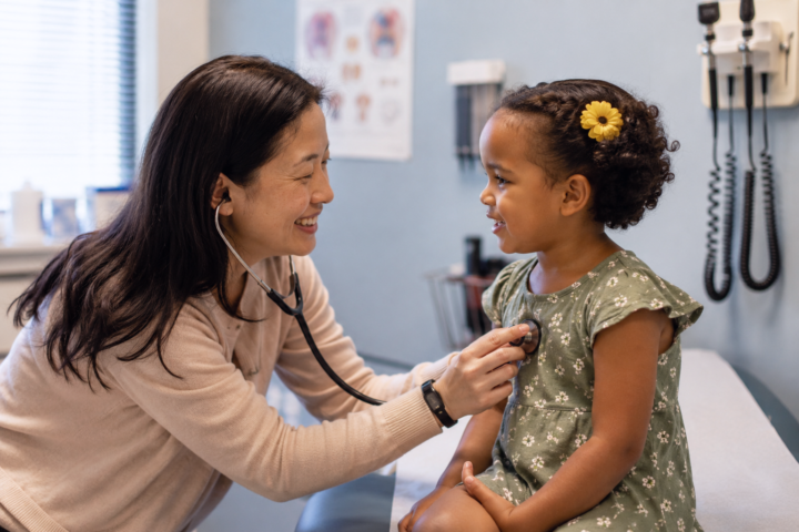 a brown haired female pediatric doctor standing with arms crossed in an exam room. the room has a picture of a giraffe and a turtle on the walls