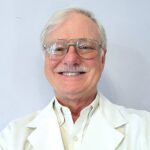portrait of a doctor with white hair and mustache smiling for hte camera. He is wearing a white coat and white shirt