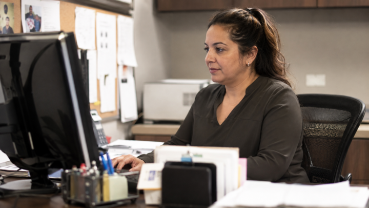 Practice administrator working at a computer in a casual clinic office, surrounded by everyday paperwork and supplies in a candid, natural setting.