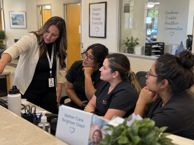 Consultant trains three front-office employees at a medical practice reception desk, gesturing toward an off-camera computer during a candid group discussion.